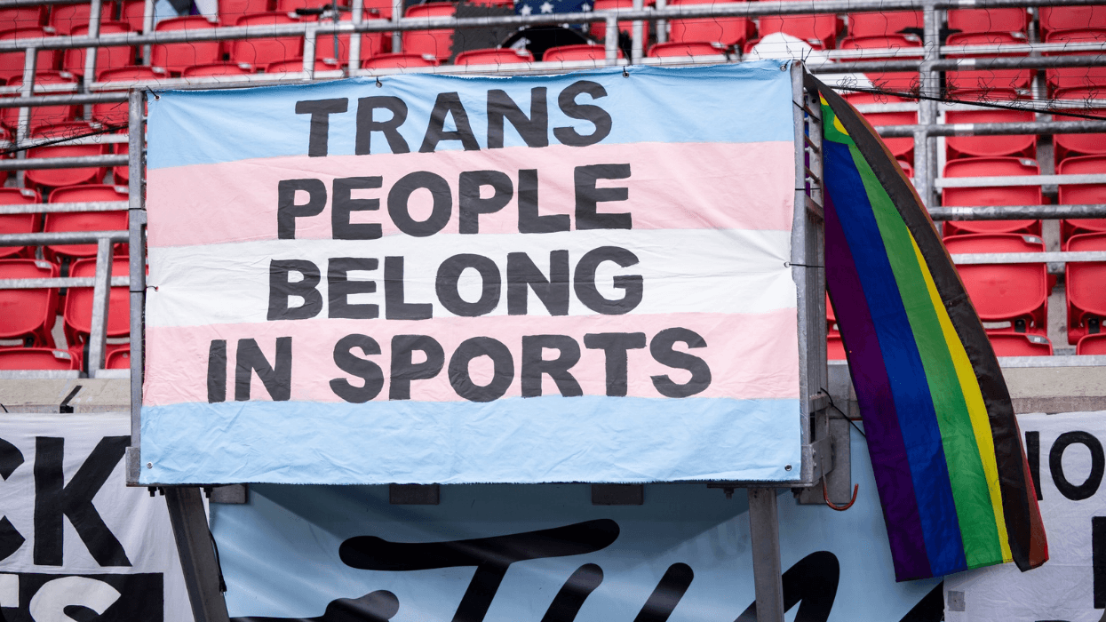 A banner with the colors of a transgender flag hangs inside of a stadium that says in all caps trans people belong in sports.