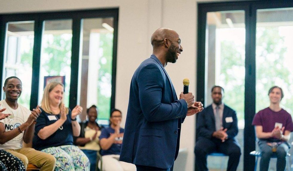a black man speaking into a microphone as he's surrounded by supporters who are clapping while seated