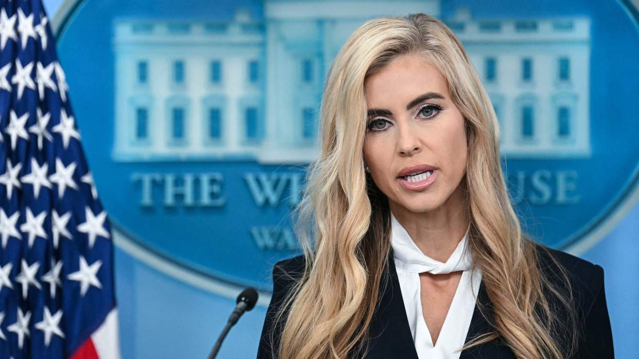 A blonde woman speaks into a microphone at a podium. She stands beside the American flag and in front of a crest of the White House.