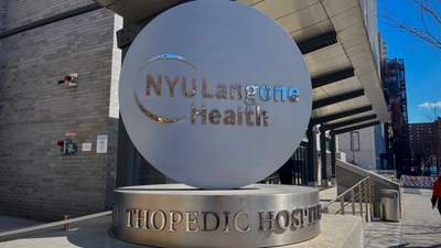 A circular silver sign on a large silver pedestal reads "NYU Langone Health Orthopedic Hospital." It is placed outside of a building on a city street.