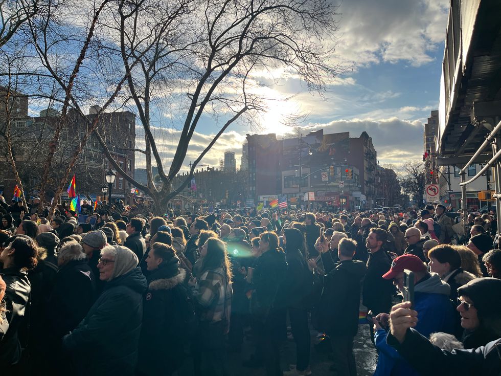 A crowd of people look left, watching something off screen. Many raise rainbow LGBTQ Pride flags.