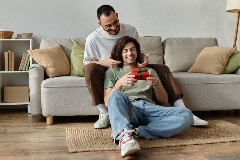 A gay man sitting on a couch who is giving his boyfriend a gift while he sits on the floor in front of him