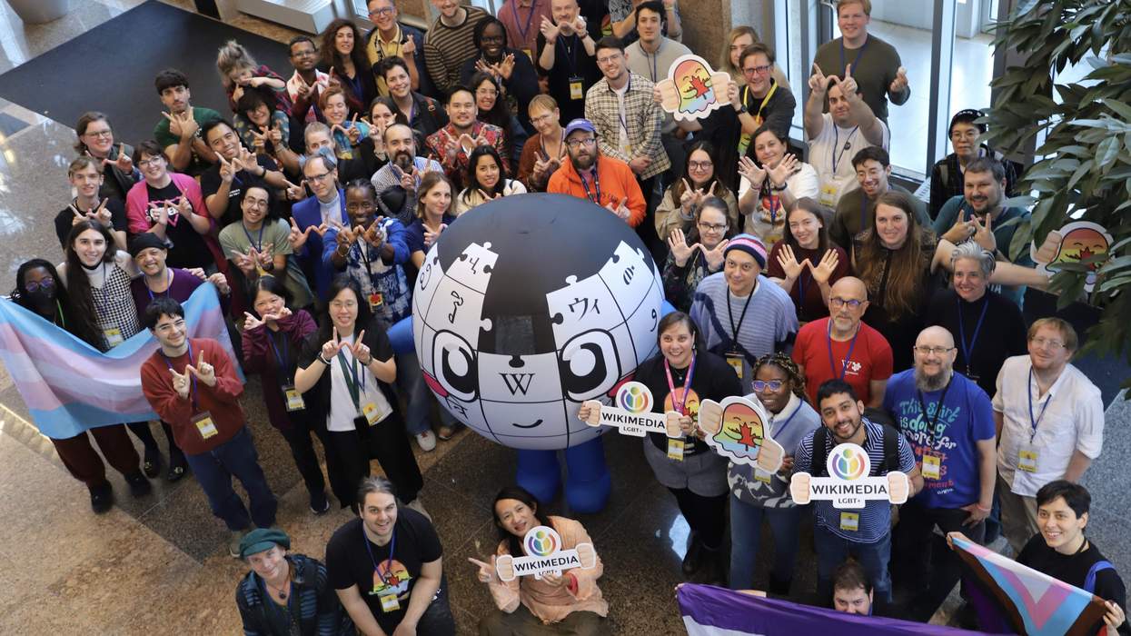 A group of people holding up LGBTQ+ Pride flags and rainbow memorabilia gather for a group photo. As the center of the group is someone dressed in a large globe costume, portraying the mascot of Wikipedia.