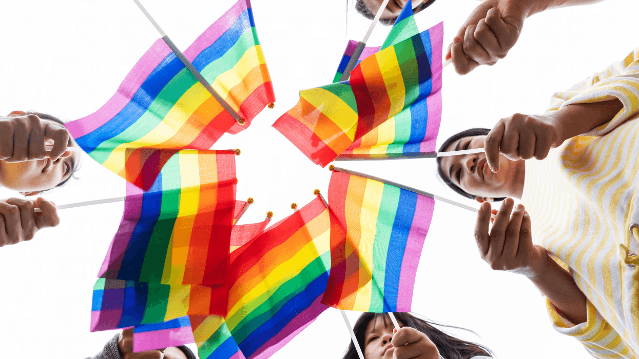 A group of people holds rainbow Pride flags together in a circle, photographed from below against a white sky.