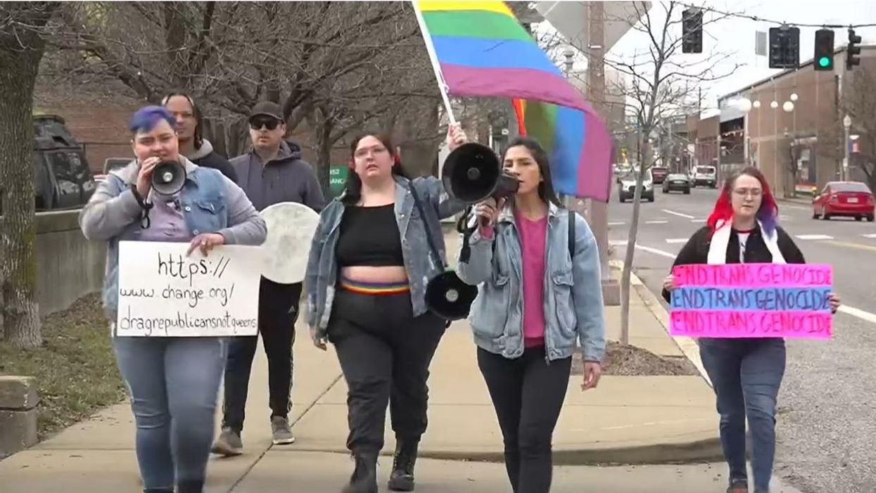 A group of people protesting while holding gay pride flags and a sign in the trans pride colors that reads "End trans genocide"