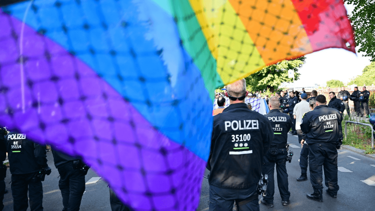 A group of police officers wearing jackets identifying them as such stand while observing demonstrators as a rainbow Pride flag hangs before them.
