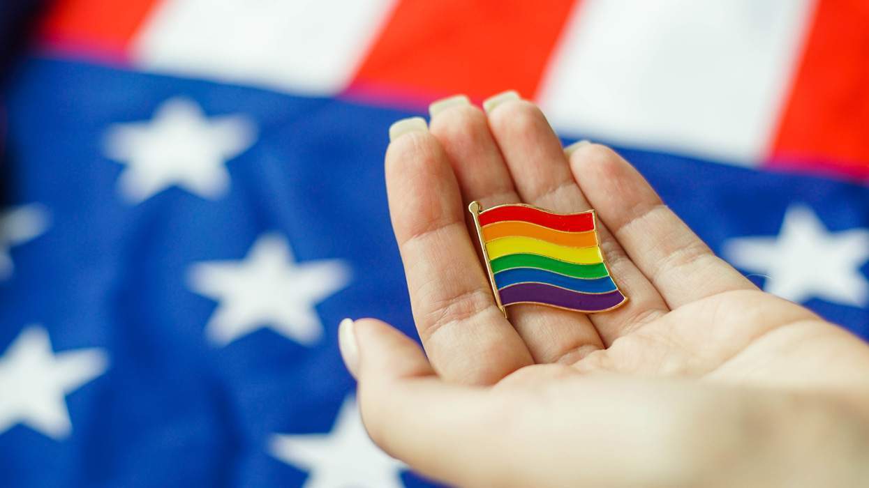 a hand holds a rainbow lgbtq pin with an american flag in the background