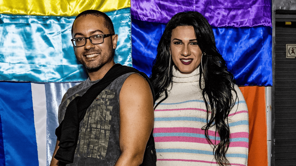 A man and a woman smile in front of a rainbow-colored backdrop.
