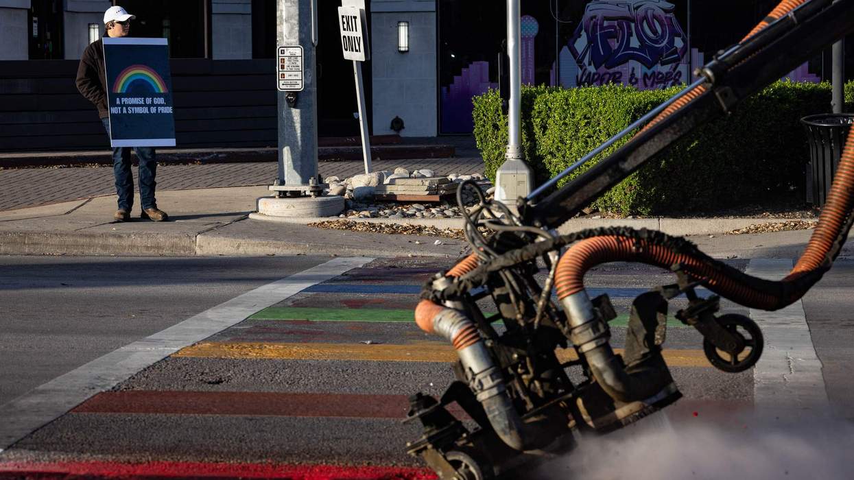 A man holds a sign with a rainbow that says A promise of God, not a symbol of PRIDE as a road crew works to scrub rainbow colored crosswalk bars off intersections.