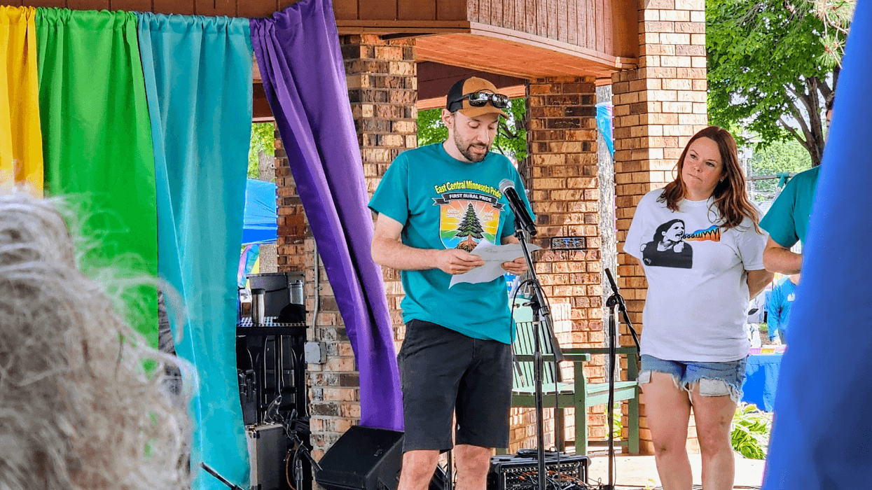 A man in a hat and sunglasses stands at a microphone and reads from a piece of paper. Behind him is a rainbow banner.