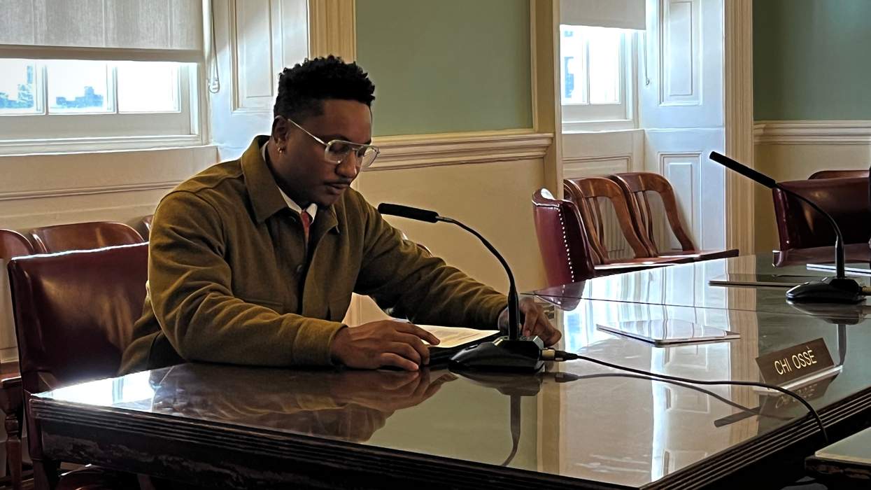A man in a quarter-zip swearer and tie sits at a desk and speaks into a microphone, reading from a sheet of paper. He sits behind a placard that reads "Chi Ossé."