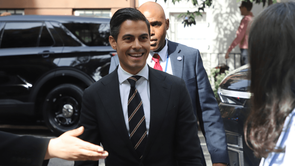 A man in a suit and tie approaches a person on a city street. He is followed by security personnel.