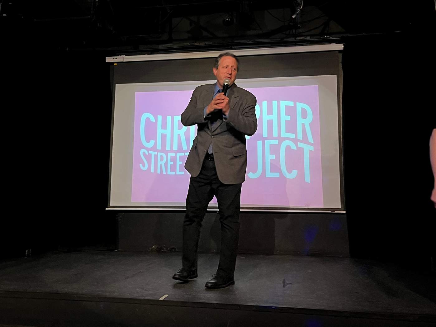 A man in a suit stands on a stage and speaks into a microphone. Behind him, a projector displays the words "Christopher Street Project."