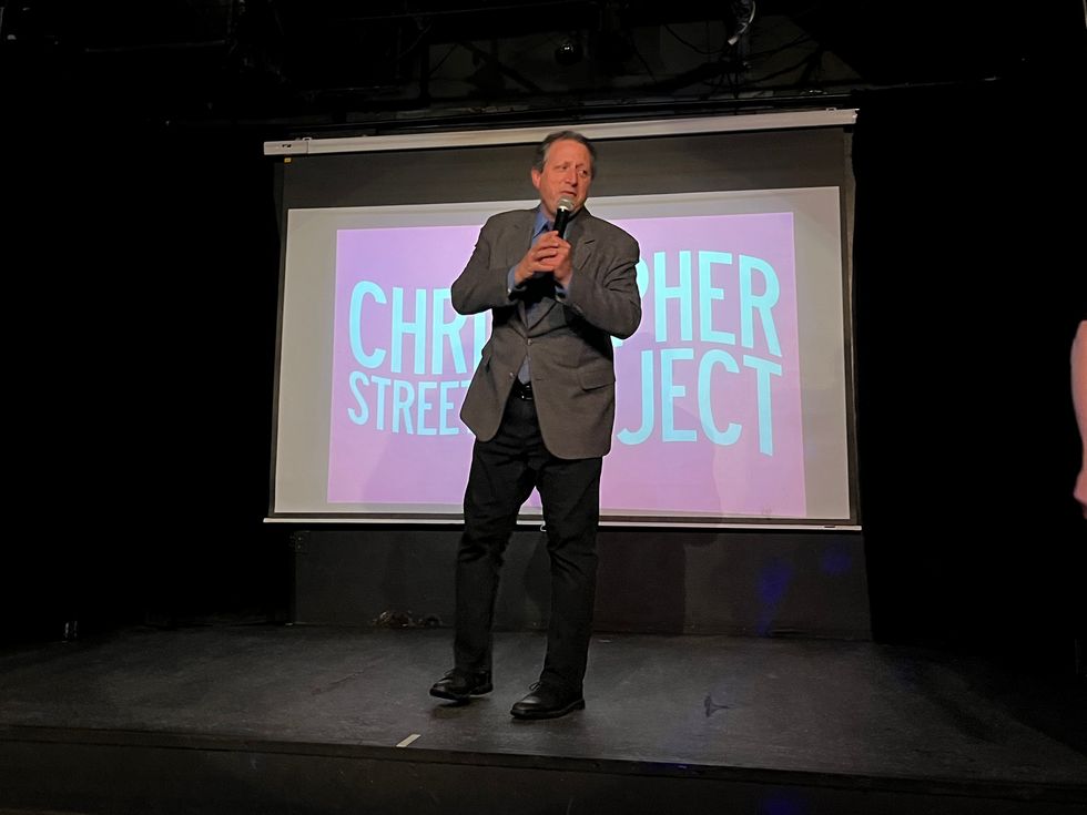 A man in a suit stands on a stage and speaks into a microphone. Behind him, a projector displays the words "Christopher Street Project."