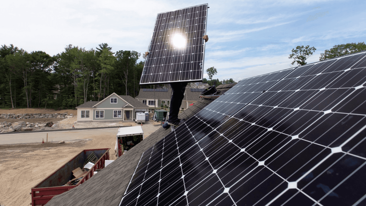 A man is standing on the roof of a home, partially covered by holding a solar panel.