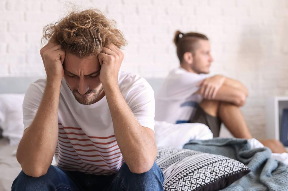 A man sitting on a bed holding his head while another man looks off into the distance.