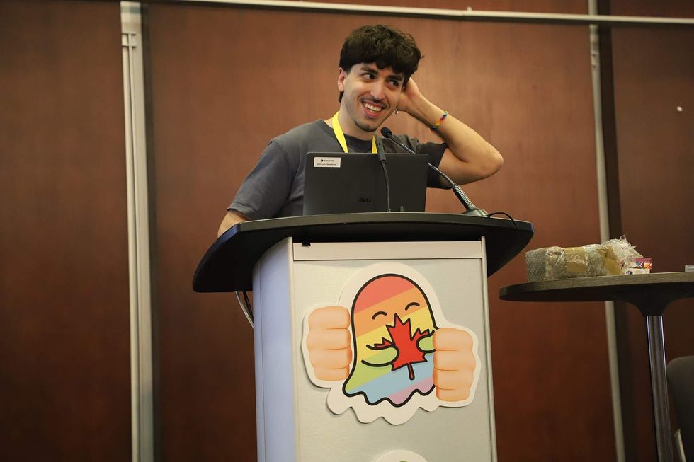 A man speaks into a microphone at a podium that displays a rainbow-colored ghost mascot.