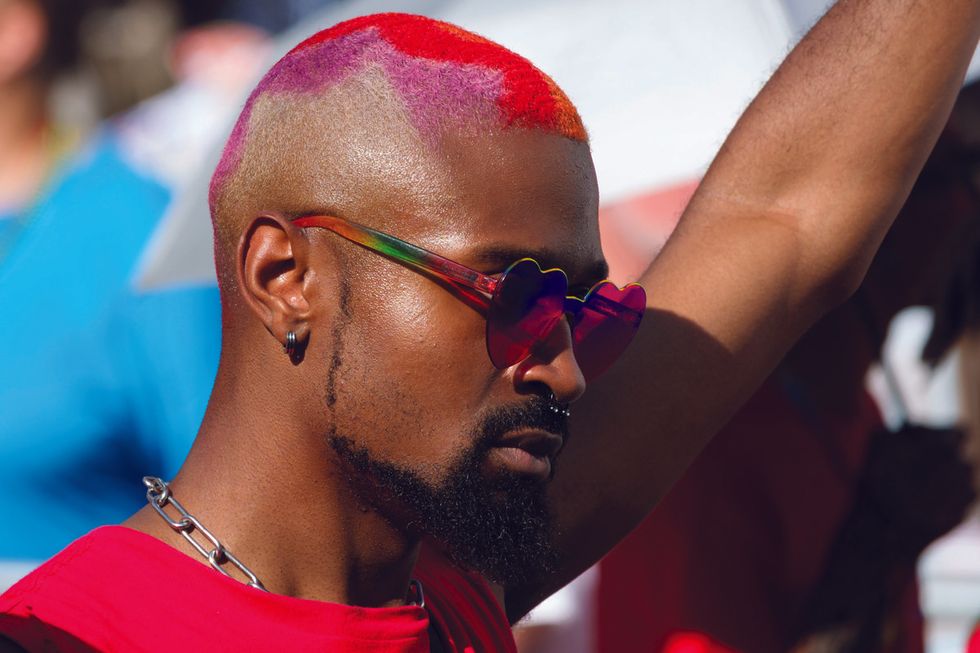 a man with pink and red hair and colorful sunglasses and red shirt raises his arm in solidarity
