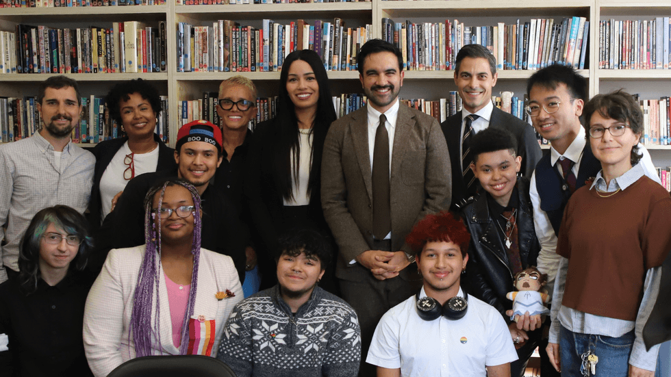 A mix of adults in formalwear and young people dressed in colorful attire stand in a library.