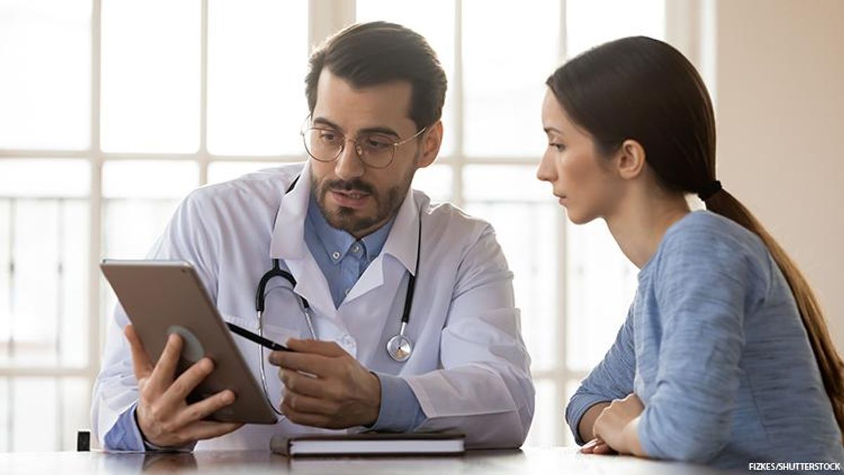 A patient looks at a clipboard that a doctor his holding while explaining something to them.