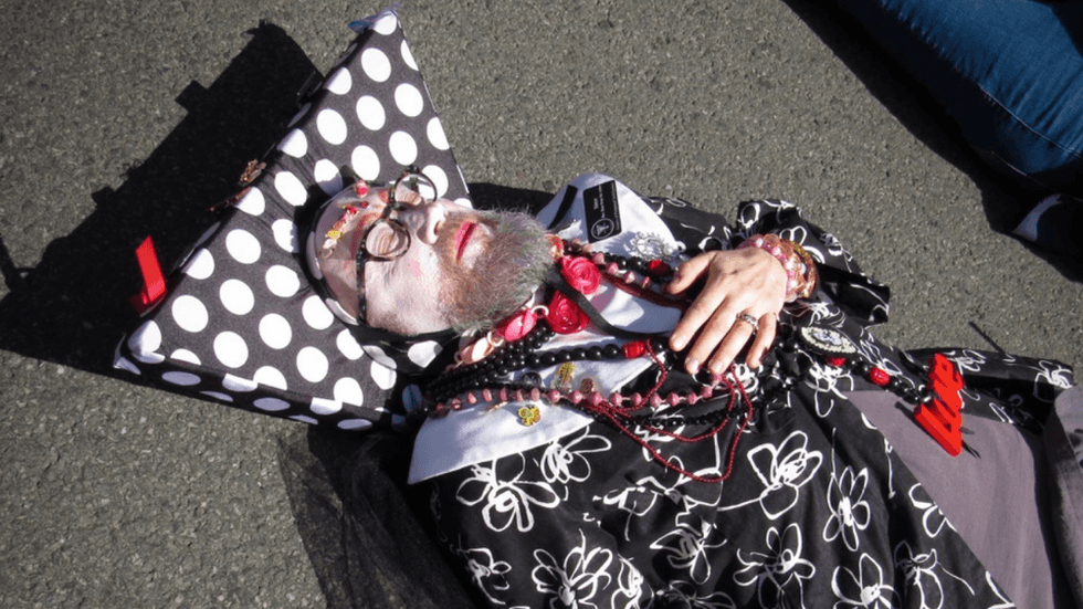 A person adorned with black and white clothing and colorful jewelry lies on the pavement of a street with their eyes closed, holding a hand to their heart.