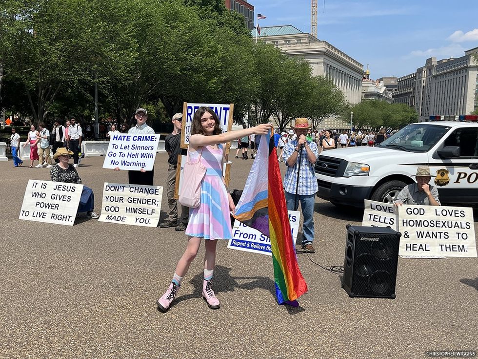 A person dressed in a trans pride flag standing in front of religious bigots with anti-LGBTQ+ signs