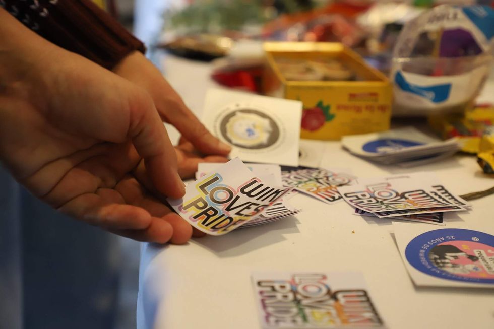 A person grabs a sticker off a crowded table. It reads "Wiki Loves Pride" overlayed on a rainbow.