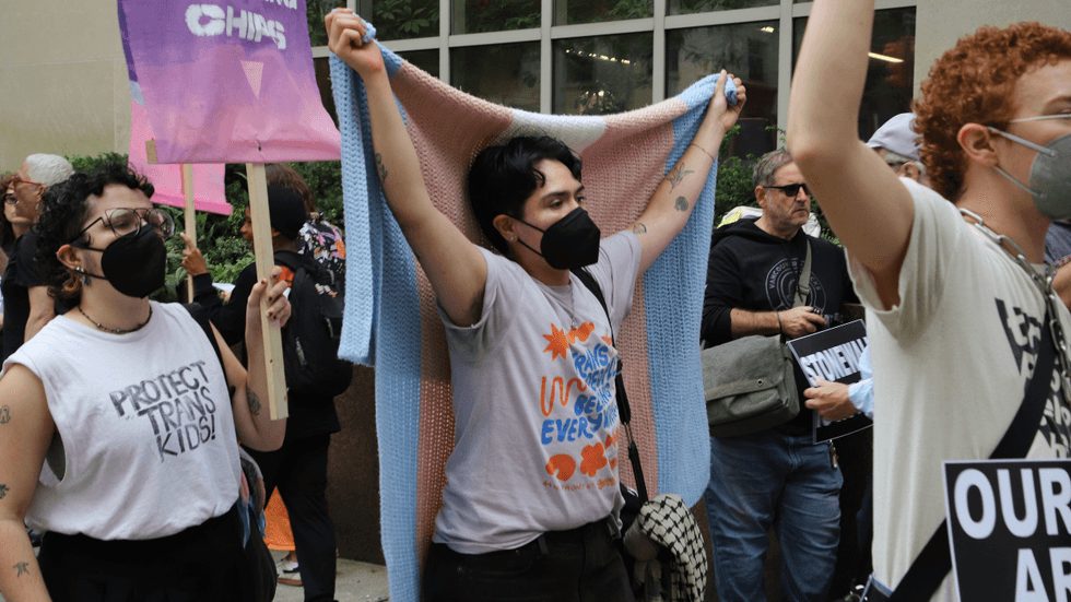 A person holds up a knit blanket bearing the blue, pink and white trans flag.