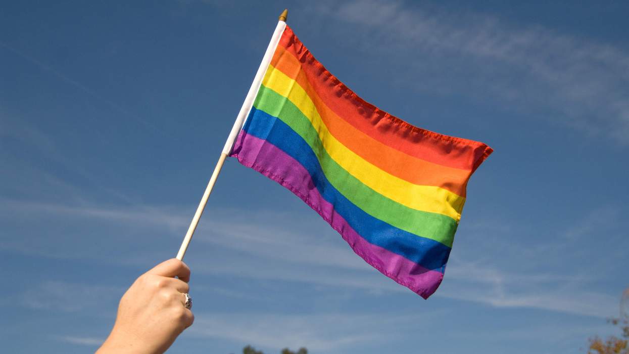 A person holds up a Pride flag toward the sky. Only their arm holding up the flag is visible in the photo.