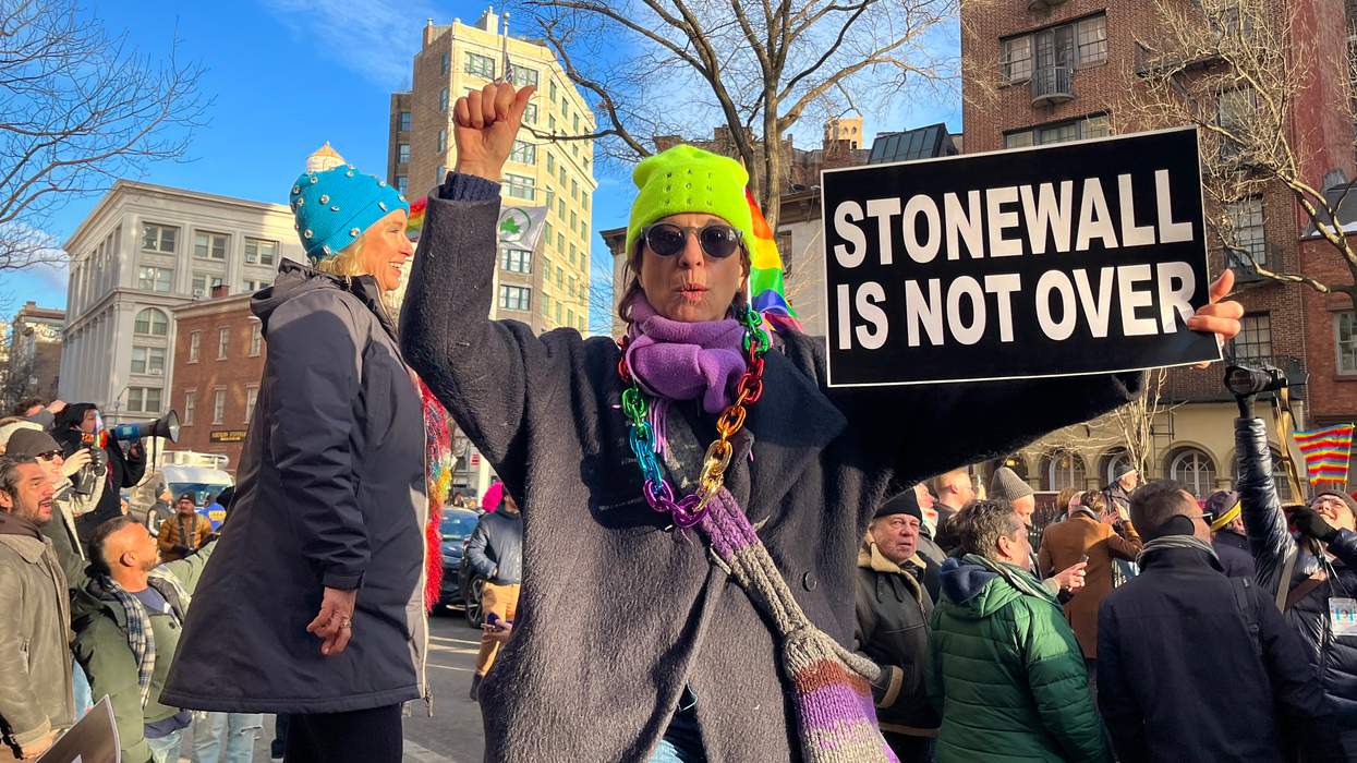 A person in a black jacket and rainbow accessories holds up a sign that reads "Stonewall Is Not Over" and raises their opposite fist in the air.