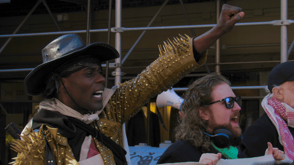 A person wearing a gold, spiky jacket and black cowboy hat raises a fist in the air and appears to be calling out something.