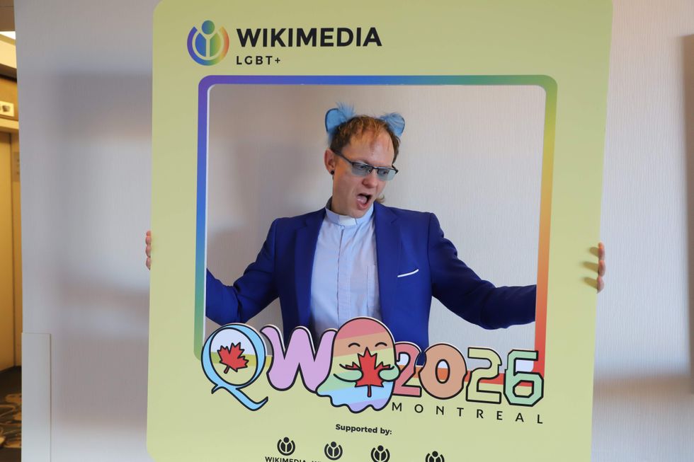A person wearing all blue clothing and cat ears holds up a yellow cardboard frame with a rainbow-colored ghost and text that reads
