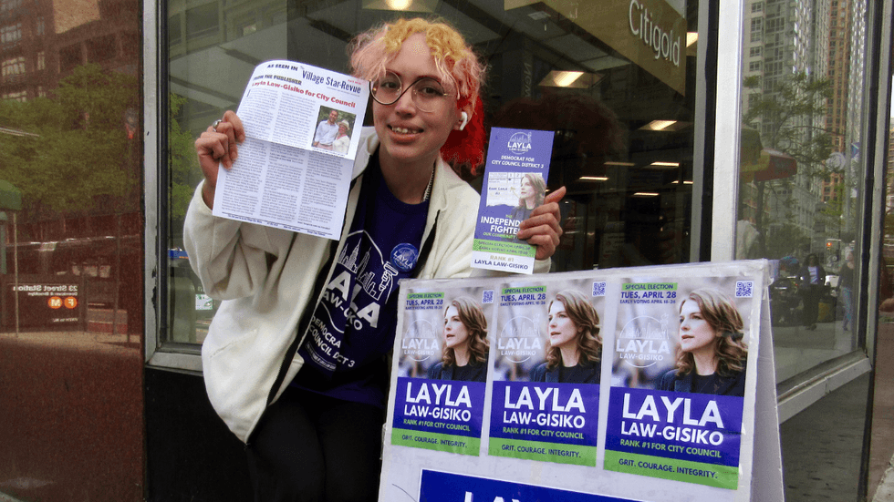 A person with multicolor hair holds up campaign handouts that read "Layla Law-Gisiko." They are standing on a city street corner.