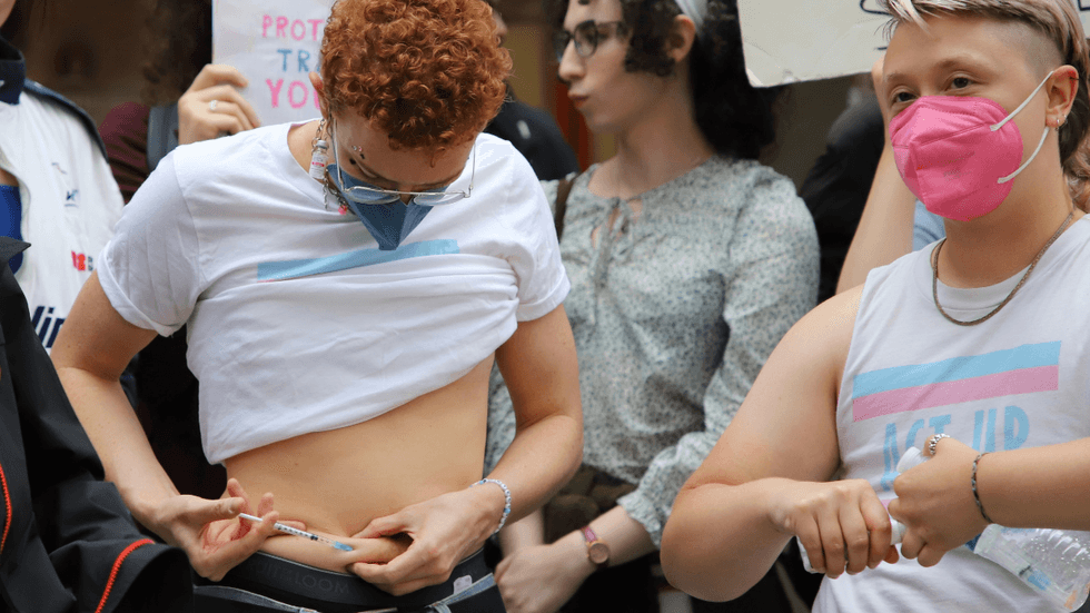 A person with red hair and a blue face mask pulls up their shirt and pokes a needle into the skin of their stomach.