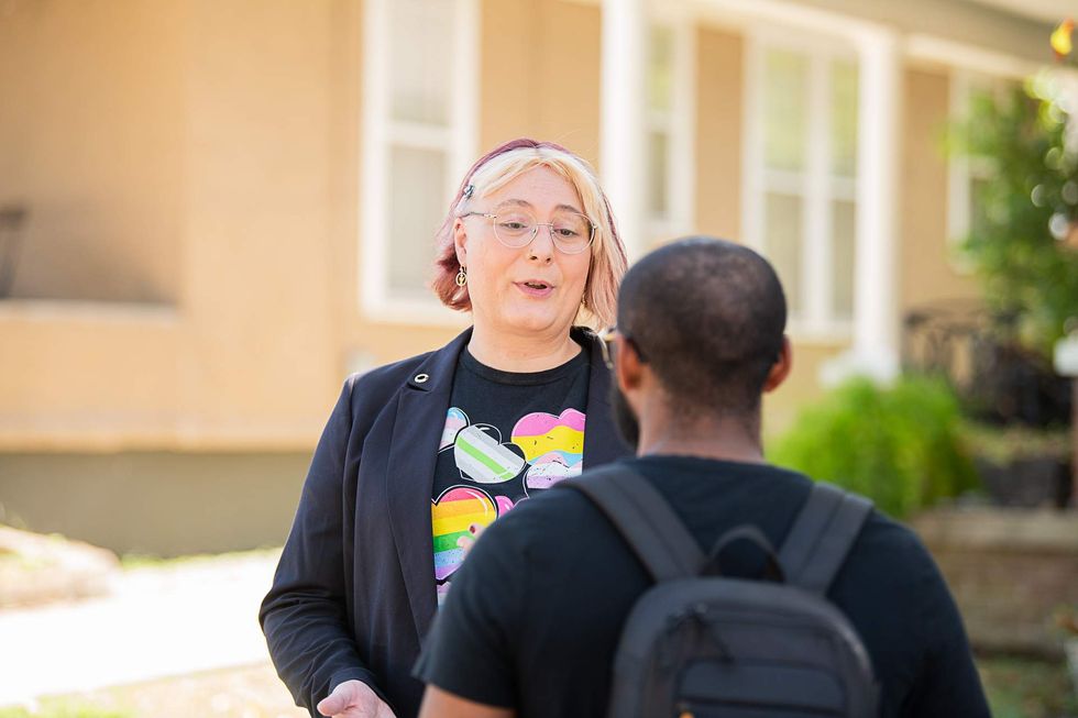 A person with short, multicolor hair and a shirt displaying several LGBTQ Pride flags speaks to a person wearing a black shirt and backpack outside.