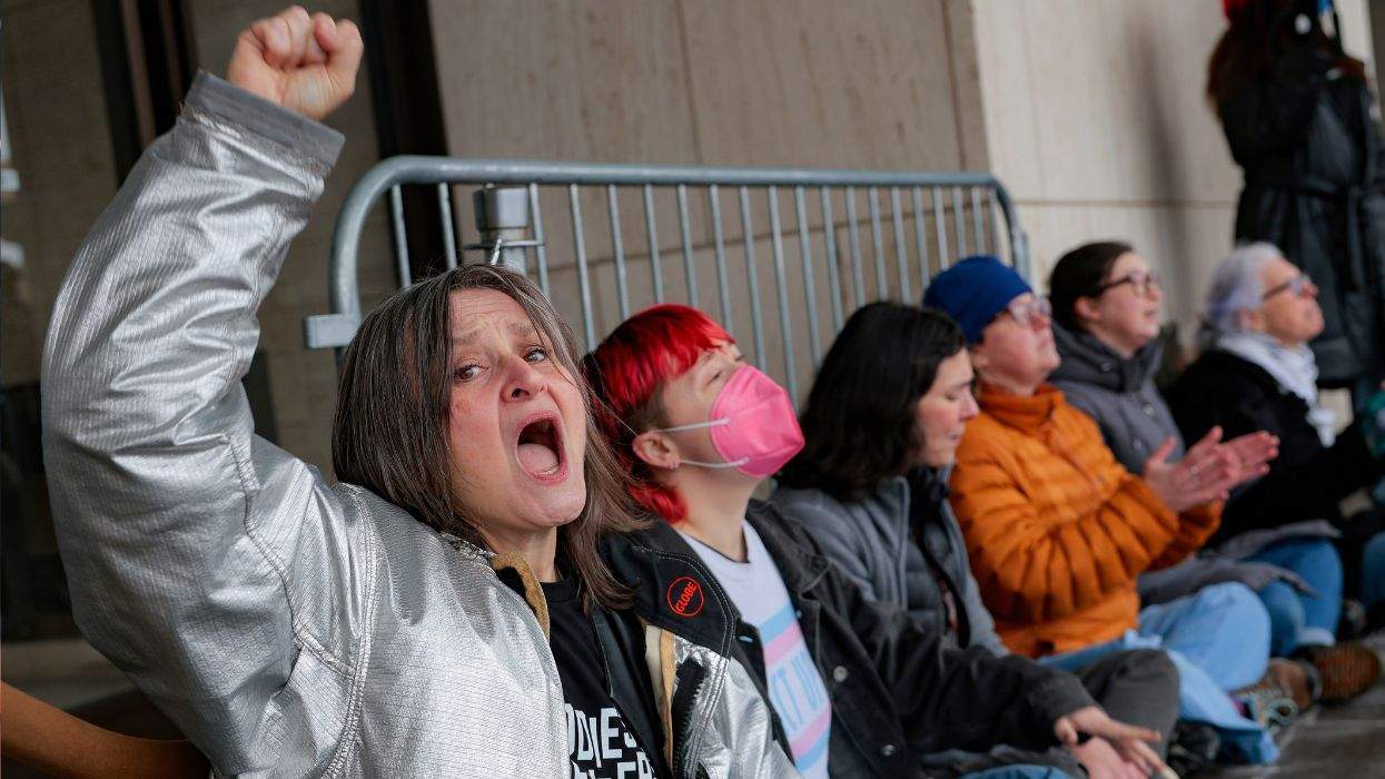 A person yells while raising their fist in the air, next to others seated on the ground in front of a building