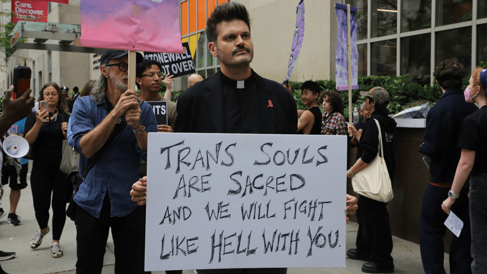 A priest with a white collar holds a sign that reads: "Trans souls are sacred and we will fight like hell with you."