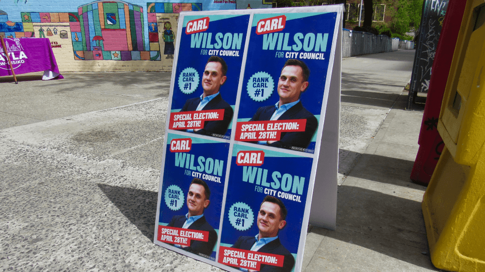 A sign standing on the sidewalk reads "Carl Wilson for the City Council."