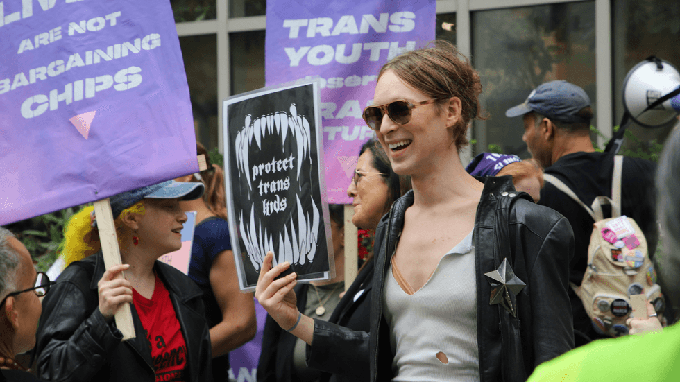 A woman holds a sign that reads: "Protect trans kids." She smiles and talks to another attendee.