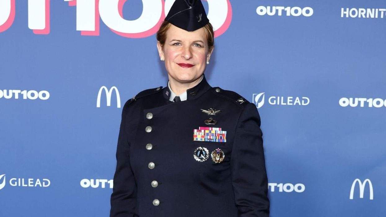 A woman in a military uniform stands before a poster that reads "Out100."