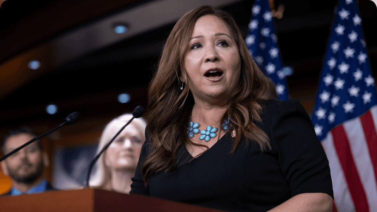 A woman in formal attire stands at a podium with microphones and speaks to an audience outside the photo.