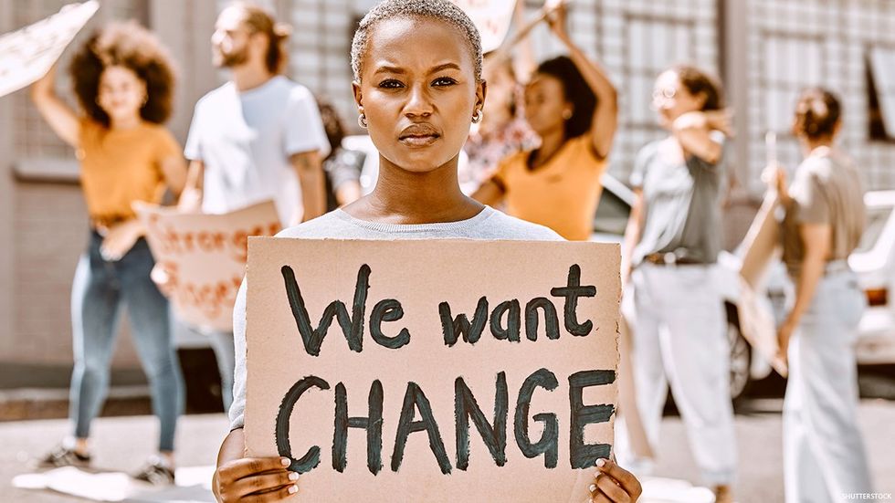 A woman stands in front of a group of people in the background holding signs. She is Black. She is holding a sign that reads "We want CHANGE."