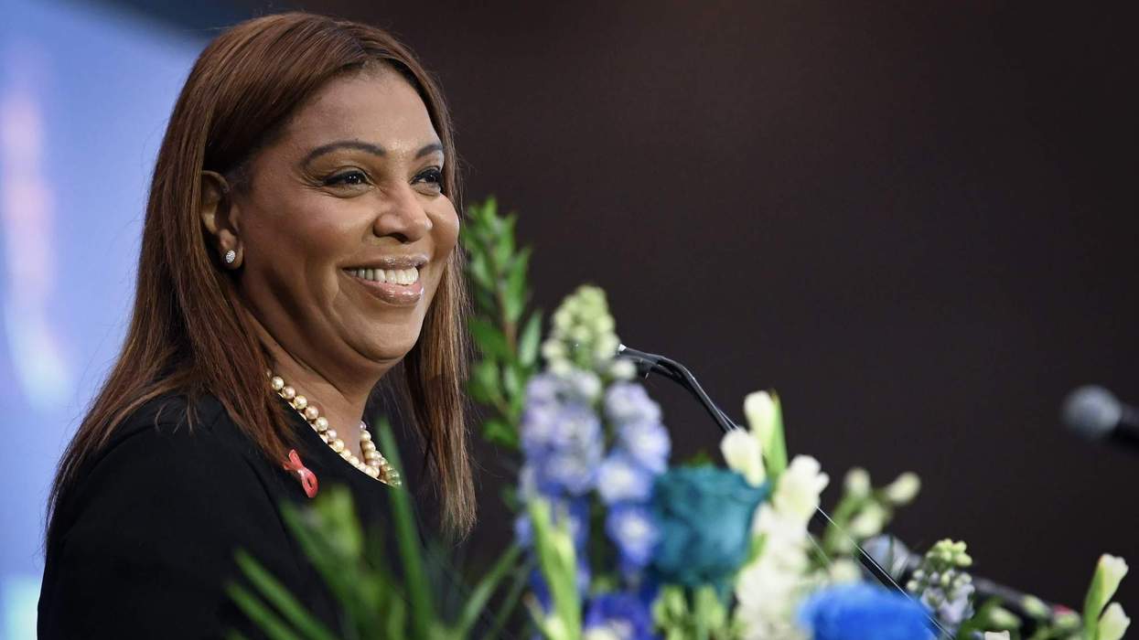 A woman with straight, brown hair and formal attire stands at a microphone smiling. She is partially covered by a bouquet of flowers.