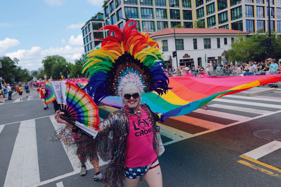 an activist wearing a colorful outfit and feather headdress helps carry a large rainbow flag