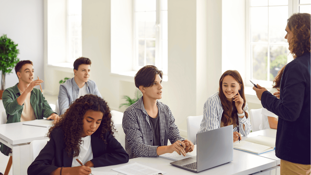 An educator speaks to a group of older students in a classroom setting while students listen, take notes, and work on laptops.