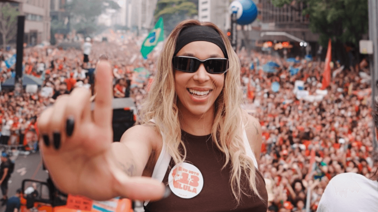 Brazilian transgender politician Erika Hilton smiles and raises her hand toward the camera while standing above a large crowd of Lula supporters waving flags at a political rally in São Paulo.