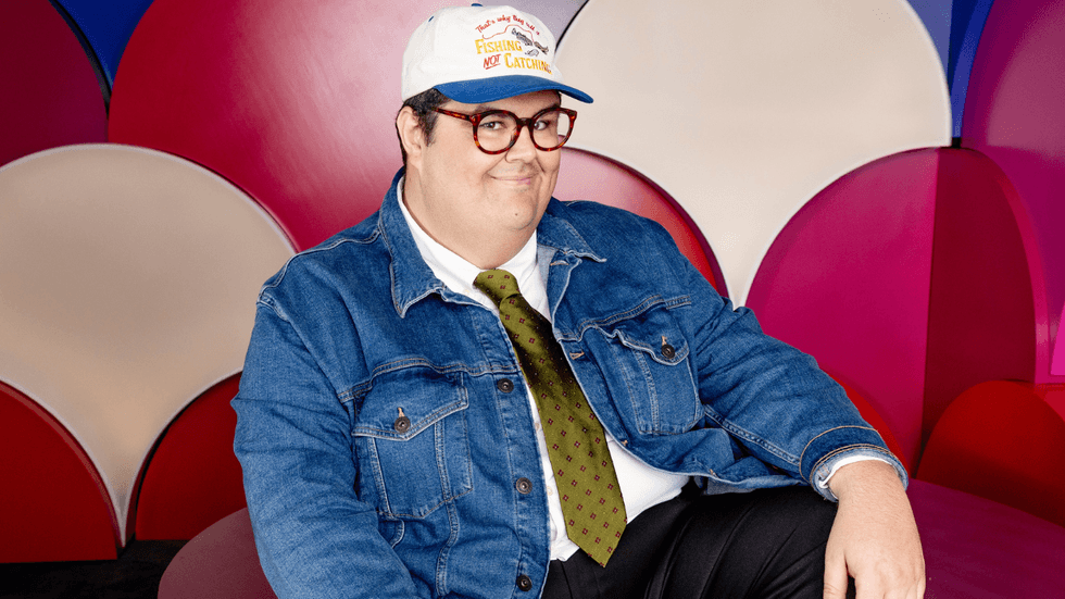 Caleb Hearon poses for the camera wearing a jean jacket, tie, hat and glasses.