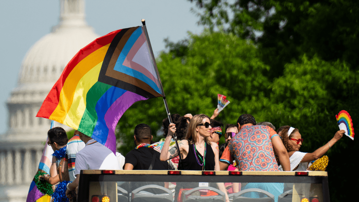 Capital Pride Parade participants wave a large Progress Pride flag from an open-top double-decker bus, with the U.S. Capitol in the background.
