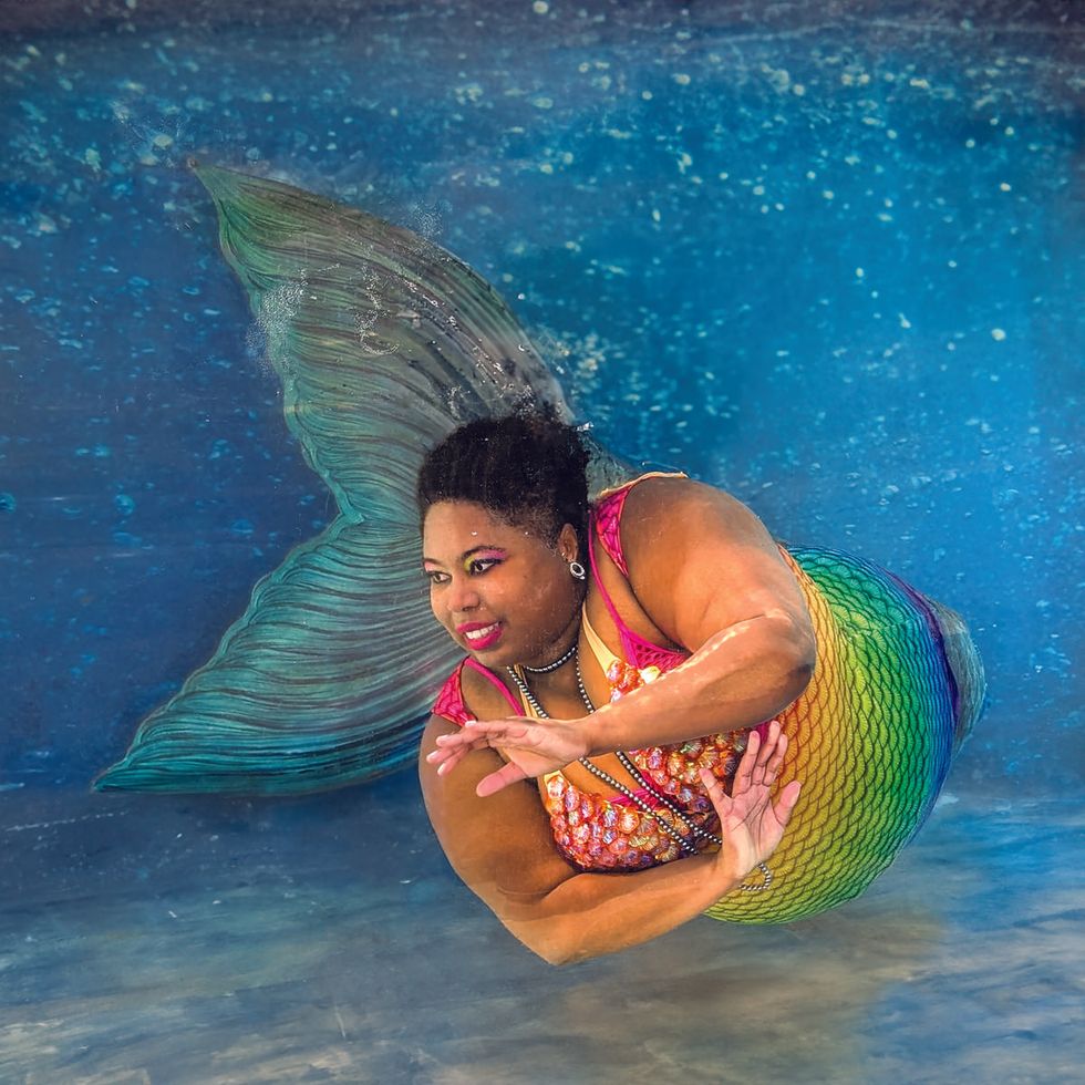 Ch\u00e8 Monique wears a colorful mermaid tail and costume while posing underwater in a swimming pool.