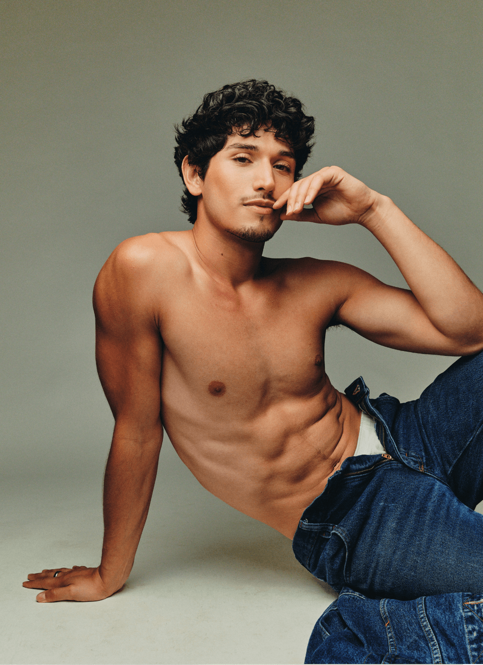 Jacob Martinez reclining shirtless in jeans, posing with one hand near his face in a studio setting